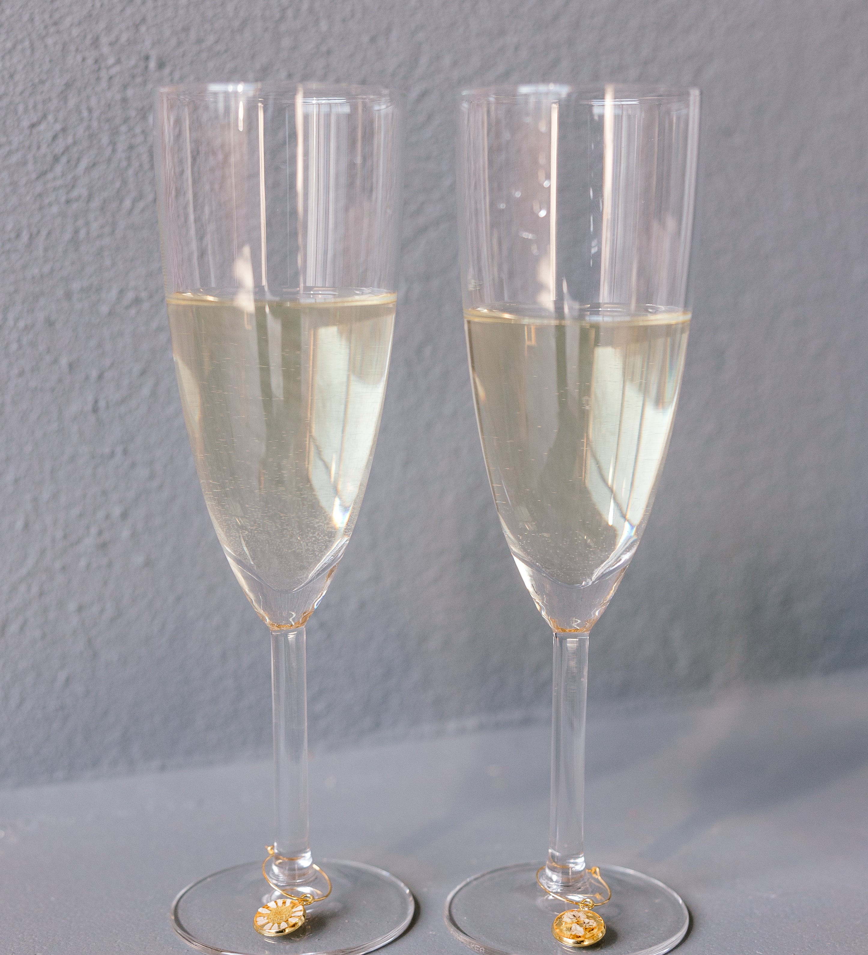 Two champagne glasses with pressed flower drink charms are placed next to each other against a grey background. The charms include real flowers. 
