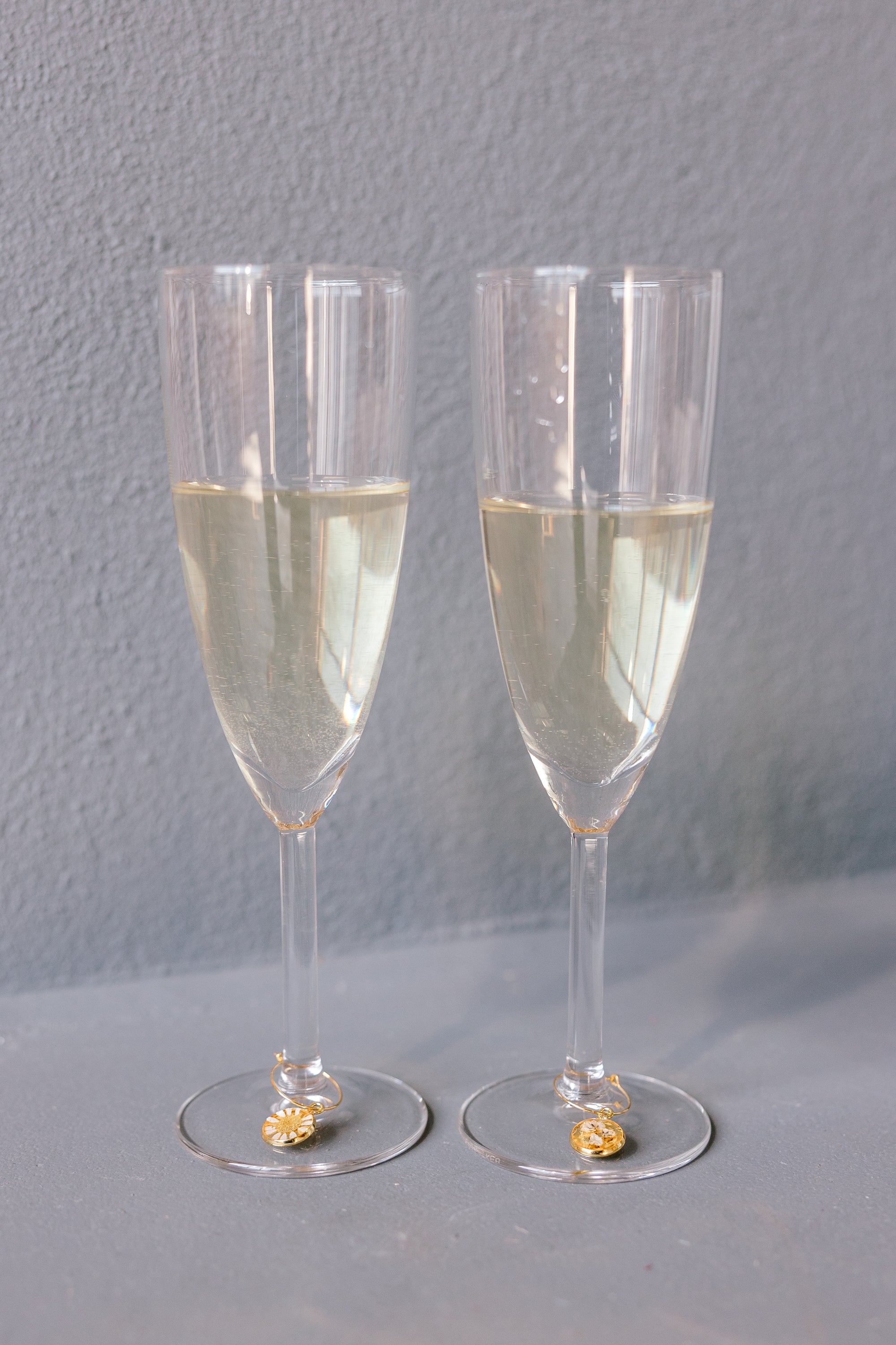 Two champagne glasses with pressed flower drink charms are placed next to each other against a grey background. The charms include real flowers. 