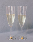 Two champagne glasses with pressed flower drink charms are placed next to each other against a grey background. The charms include real flowers. 