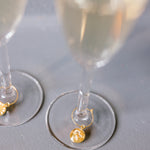 Two gold pressed flower drink charms are placed on two clear but full champagne glasses, set on top of a grey surface. 