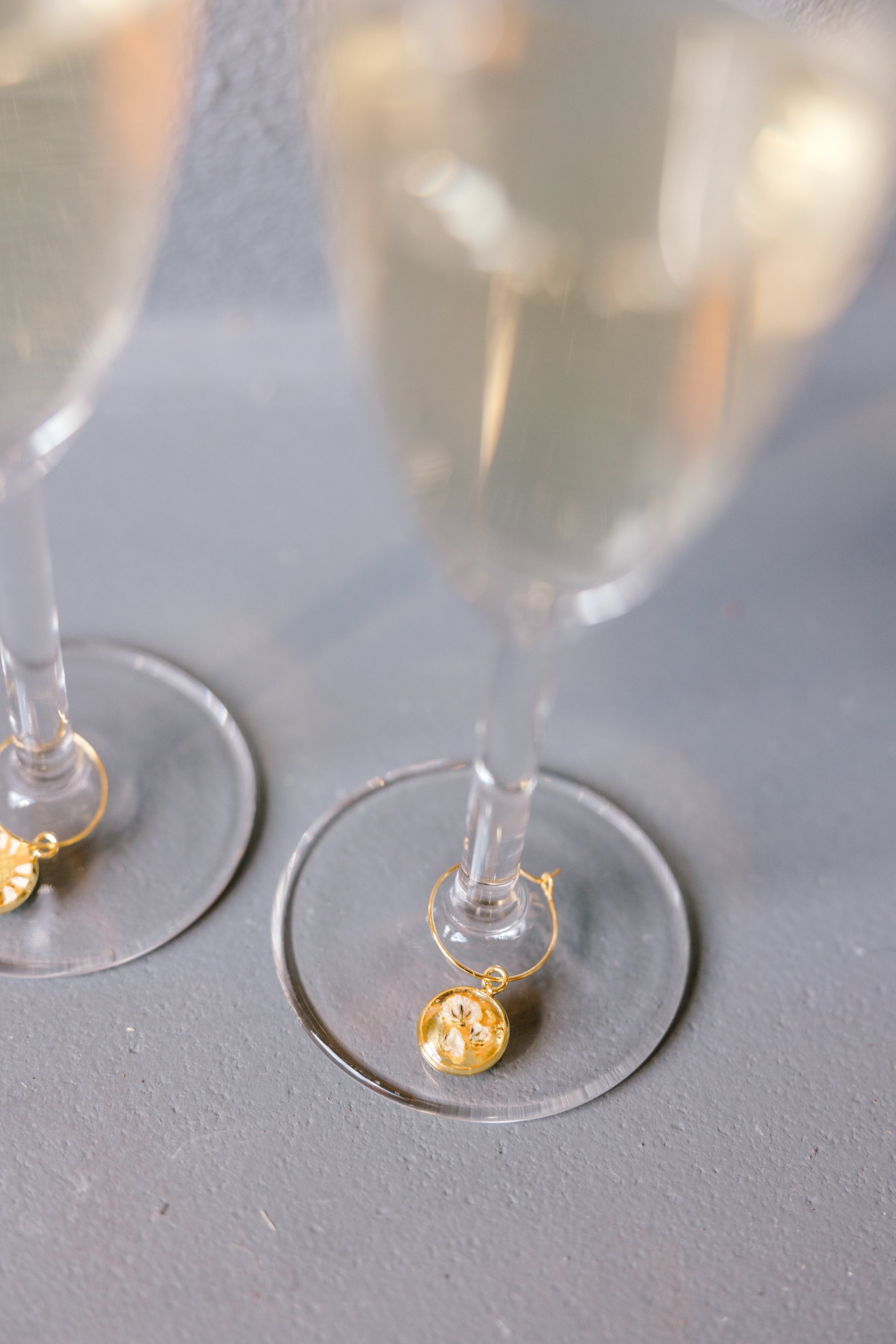 Two gold pressed flower drink charms are placed on two clear but full champagne glasses, set on top of a grey surface. 