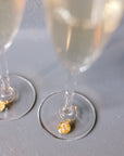 Two gold pressed flower drink charms are placed on two clear but full champagne glasses, set on top of a grey surface. 