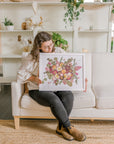 A woman sits holding her bouquet preservation she received from the Pressed Bouquet Shop. It features pink and white pressed flowers with pressed greenery on a white background and in a white frame.