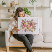 A woman sits holding her bouquet preservation she received from the Pressed Bouquet Shop. It features pink and white pressed flowers with pressed greenery on a white background and in a white frame.
