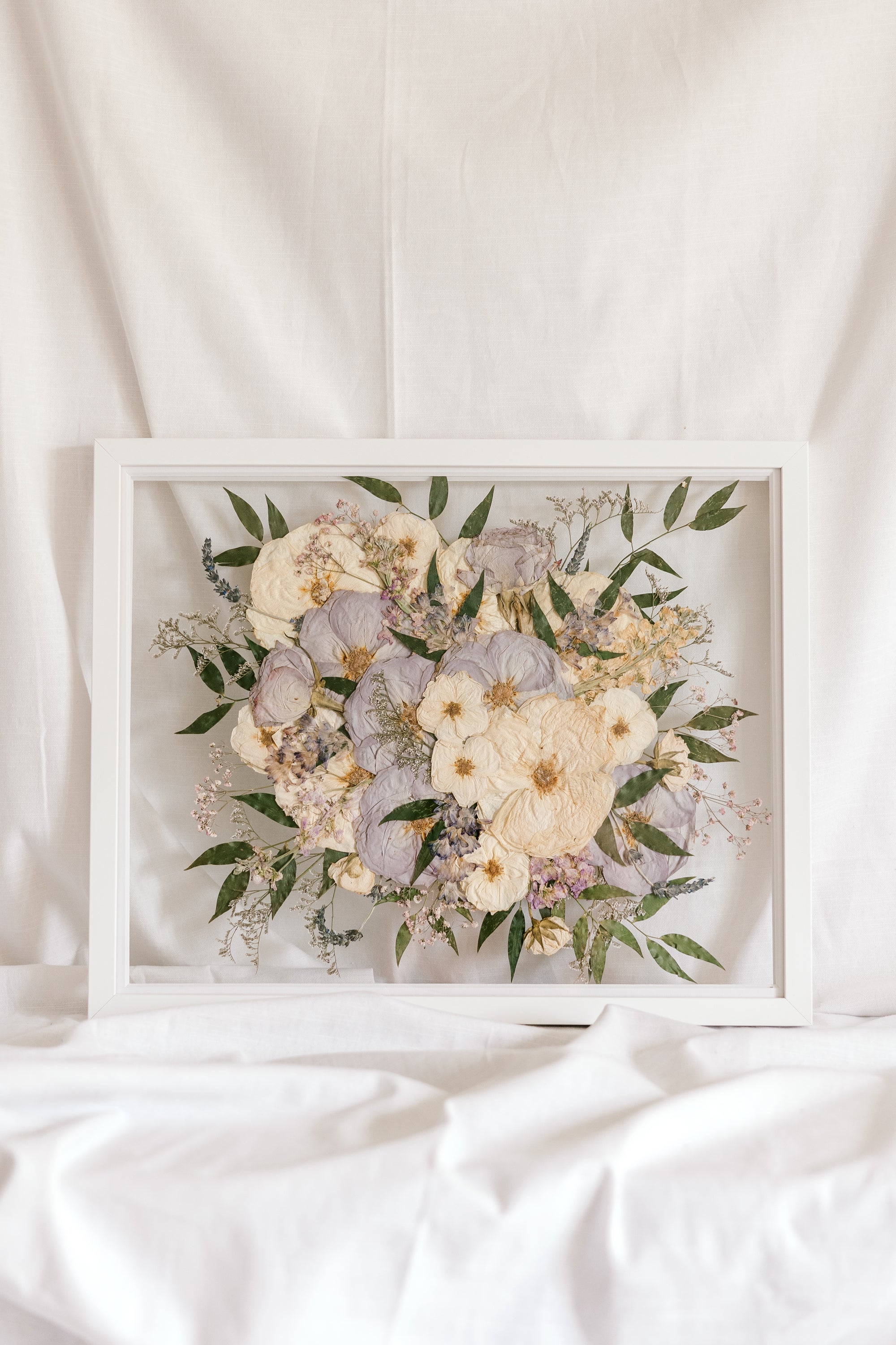 A white and light purple wedding bouquet turned into pressed flowers and displayed inside of a white wood preservation frame.