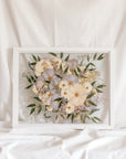 A white and light purple wedding bouquet turned into pressed flowers and displayed inside of a white wood preservation frame.