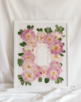 A bouquet of pressed pink peonies is displayed with a wedding invitation in a 16x20 white wood frame.