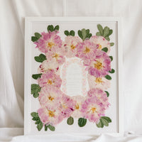 A bouquet of pressed pink peonies is displayed with a wedding invitation in a 16x20 white wood frame.