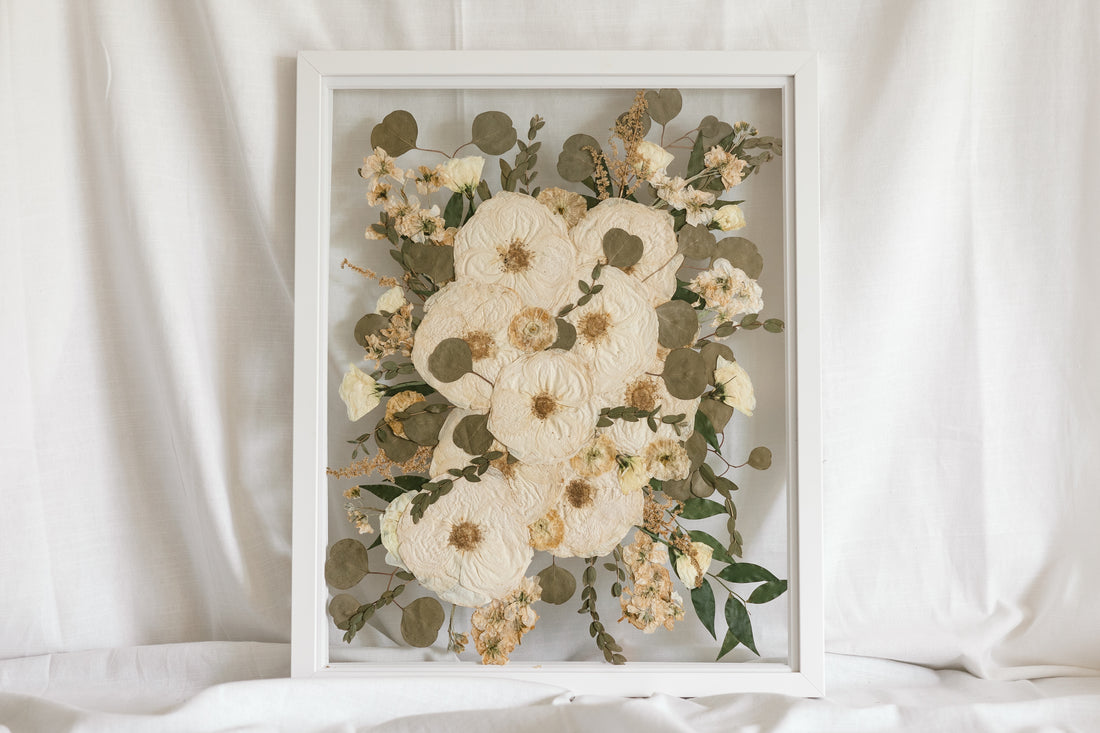 An all white and green pressed flower wedding bouquet displayed inside of a white wood glass floating frame.