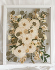 An all white and green pressed flower wedding bouquet displayed inside of a white wood glass floating frame.