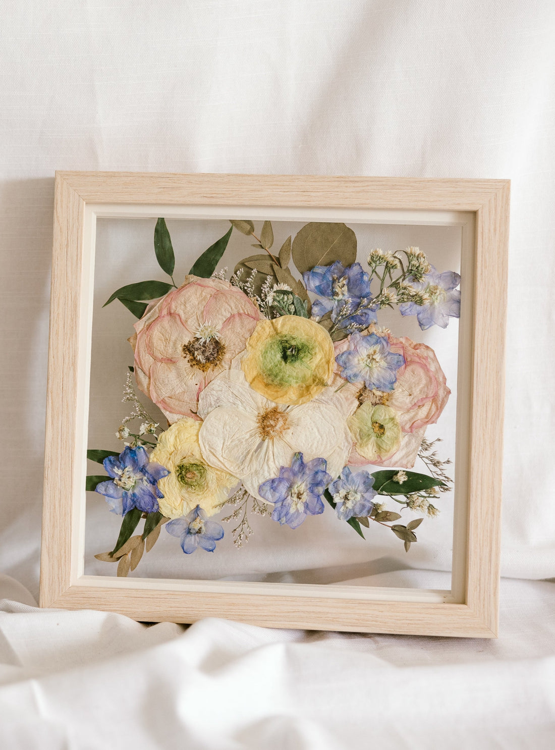 White, pink, yellow, and blue pressed flowers in a 10" square natural wood frame. 