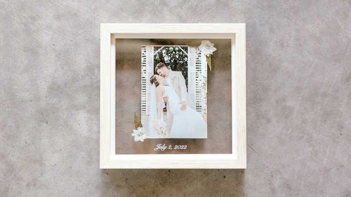 Picture of a couple that was just married holding a bouquet of flowers encased in a natural small frame with pressed flowers surrounding it.