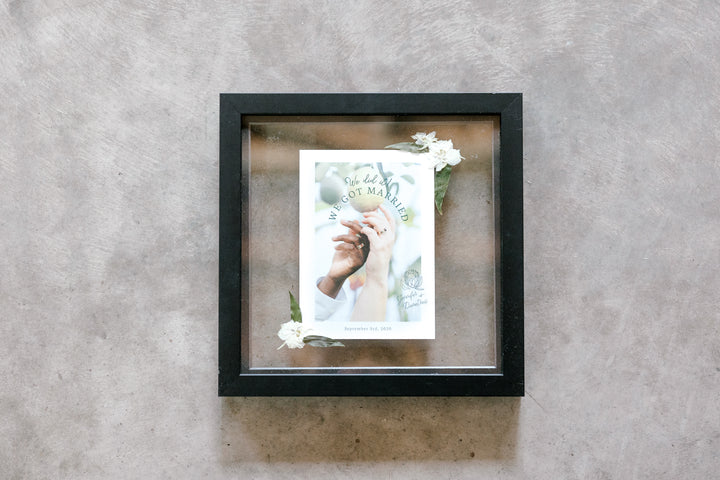 Two hands intertwined with wedding bands on in an invitation that is preserved into a small black frame.