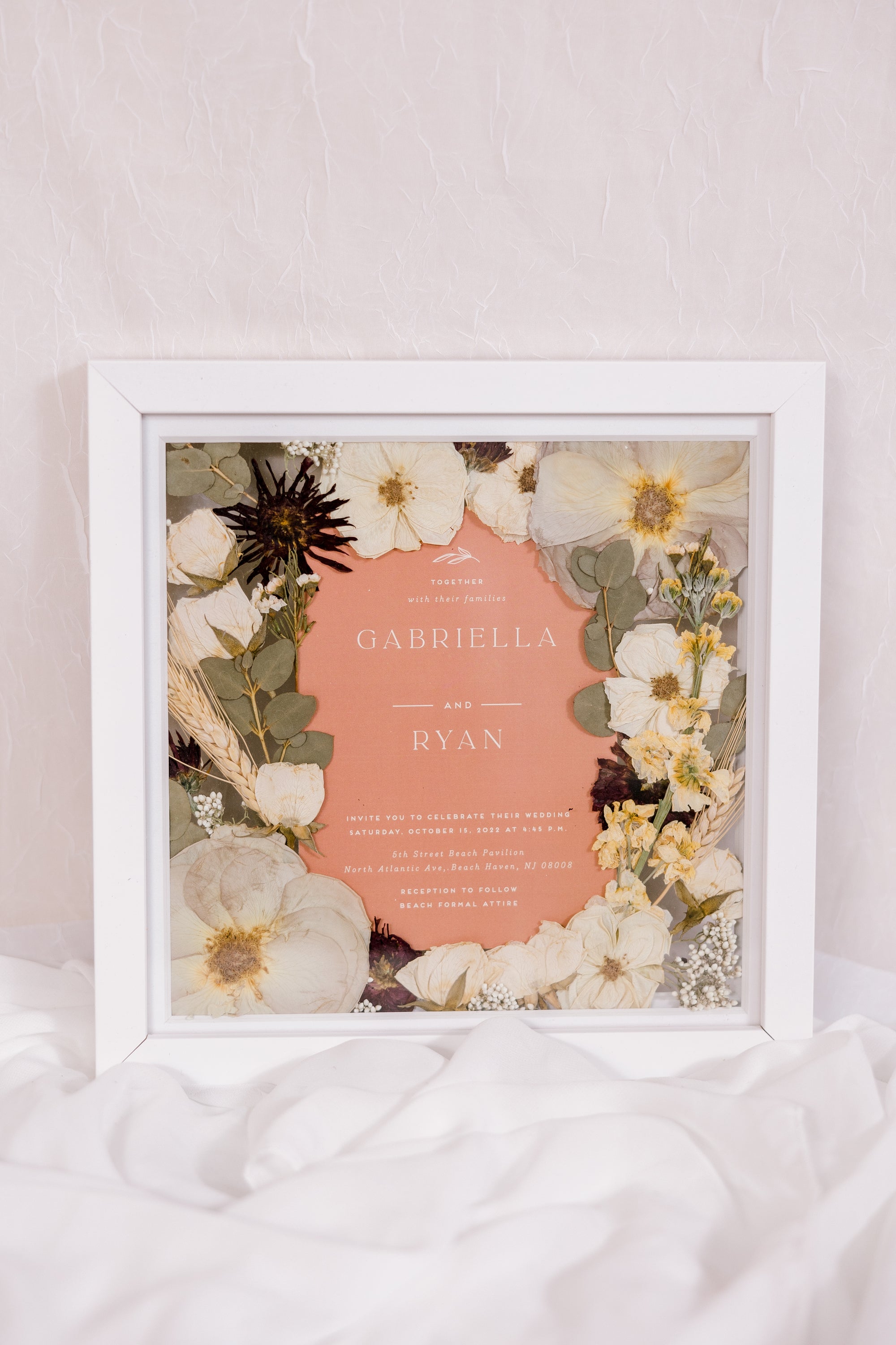 Beautifully preserved pressed flowers surround an orange-color wedding invitation in a square wood frame. 