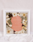 Beautifully preserved pressed flowers surround an orange-color wedding invitation in a square wood frame. 