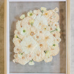 An all white ball bouquet pressed and preserved into a gold frame against concrete.