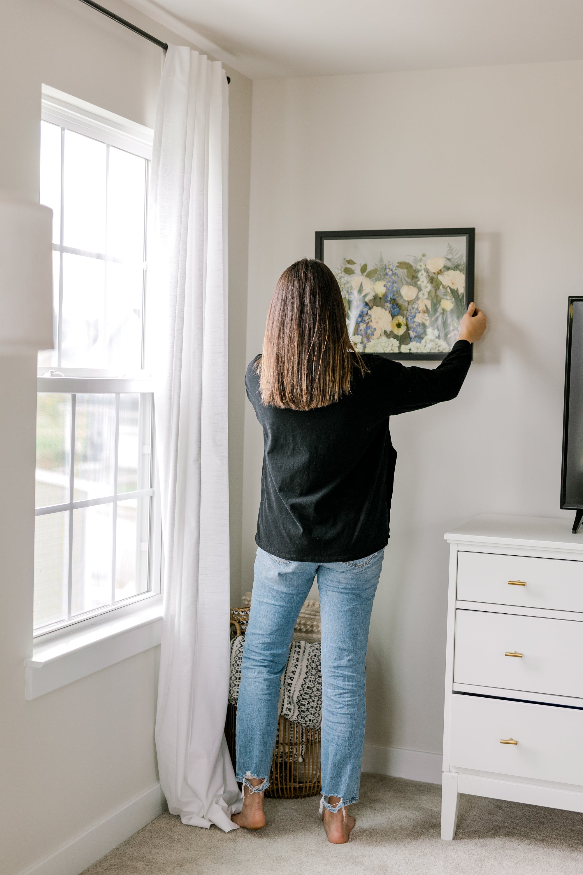 A woman stands in a bedroom hanging a frame filled with pressed flowers. 