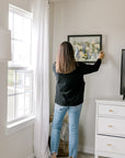 A woman stands in a bedroom hanging a frame filled with pressed flowers. 