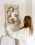 A woman hangs her pressed wedding bouquet on a wall above a blanket rack. The pressed flowers surround her wedding invitation that she smiles up at while hanging.