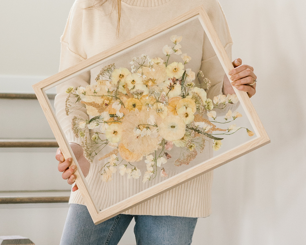 A bride is holding her pressed white bouquet, designed in a 16x20 natural wood frame with clear glass backing. The bouquet extends from the center of the frame with stems stretching to the corners.