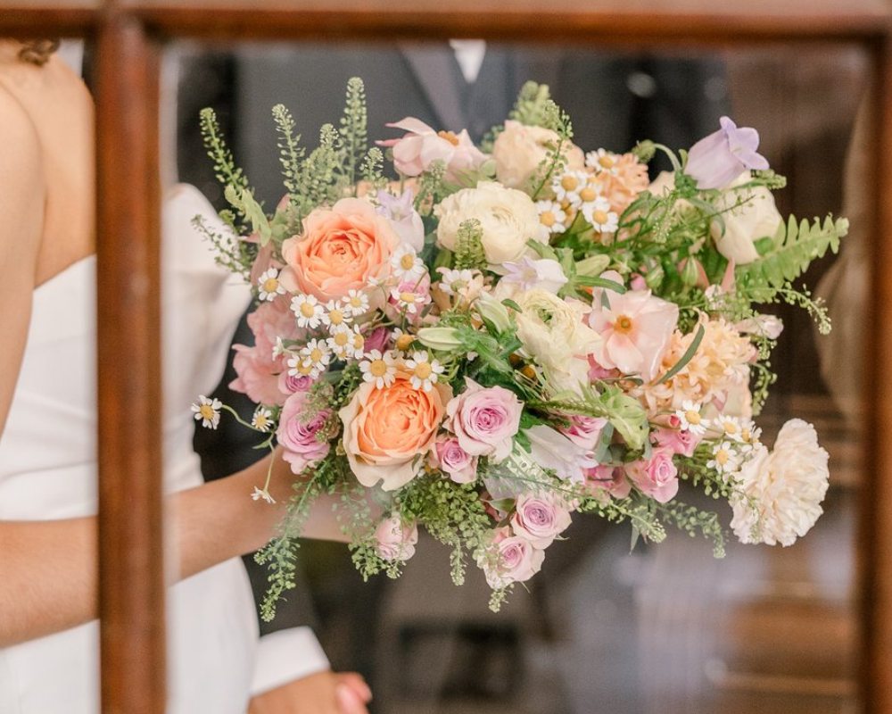 A bride is holding her wedding bouquet  in one hand and her husband's hand in the other. Her bouquet includes white, pink, and orange flowers with greenery bursting from all sides.