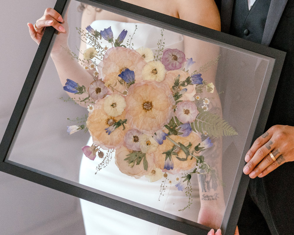 A newlywed couple is holding their pressed pink, blue, and white bouquet, preserved in a 16x20 black wood frame with a floating glass background.  