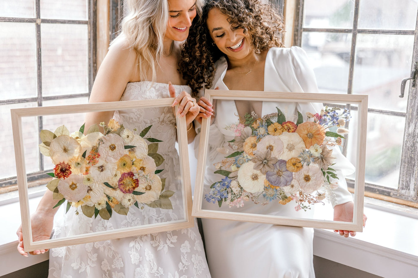 Two women stand in wedding dresses smiling down at their preserved wedding florals. 