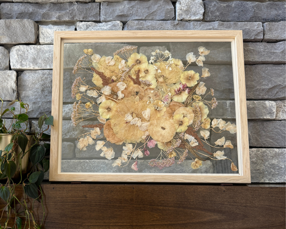 A pressed white bouquet in a 16x20 natural wood frame with glass backing is perched against a gray stone wall on a wooden mantel. 