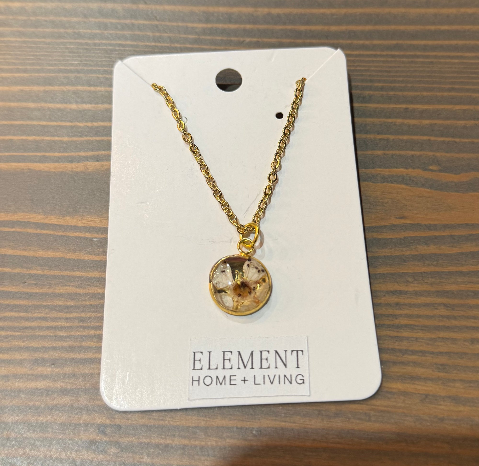 A pressed flower necklace with a tiny white flower is set in a circular gold-plated bezel cup. It is placed atop a wooden surface. 