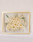 An all white bouquet of flowers pressed into a glass floating frame surrounded by gold wood.