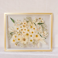 An all white bouquet of flowers pressed into a glass floating frame surrounded by gold wood.
