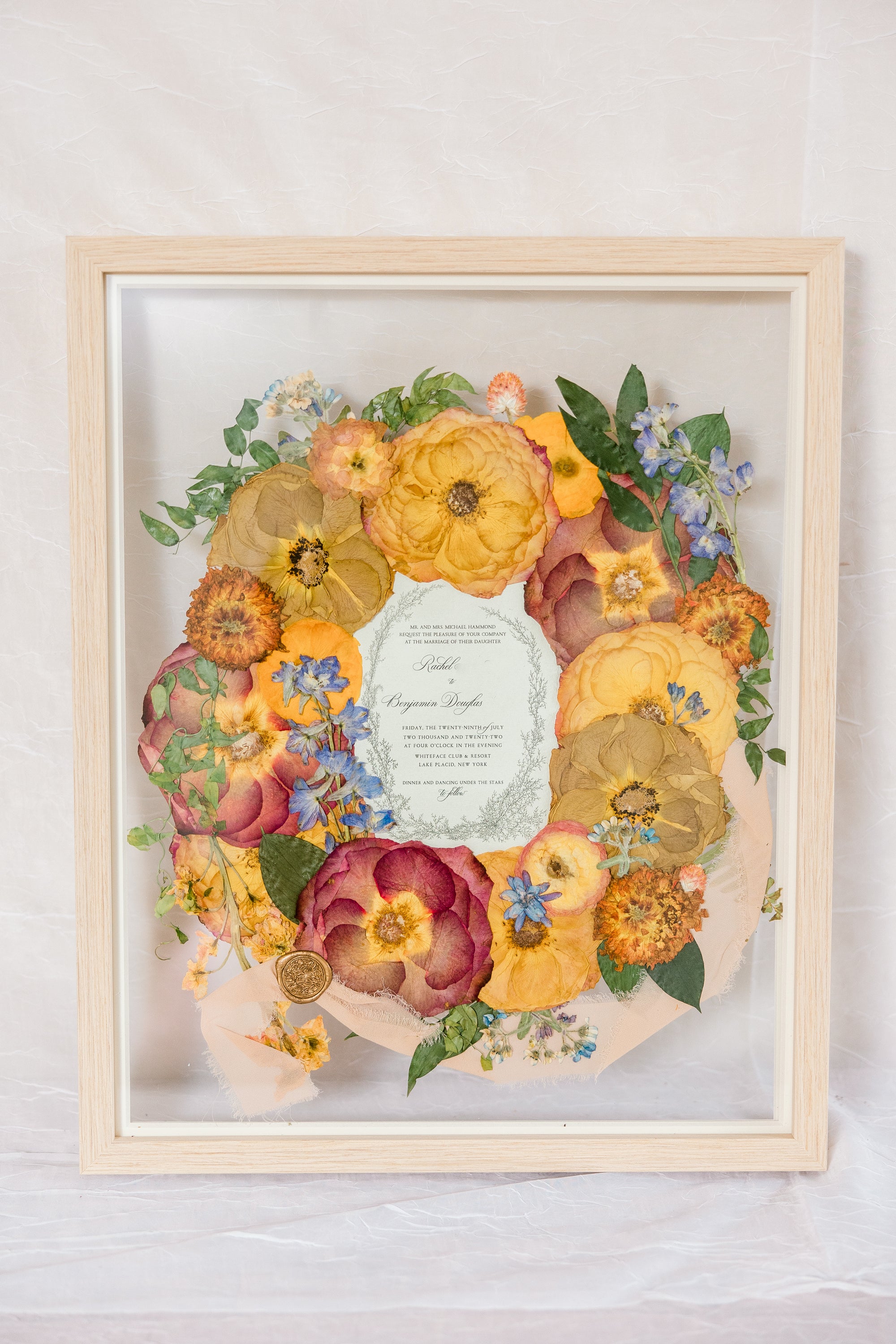 Orange and blue flowers surrounding a wedding invitation with ribbon and a wax seal, preserved in a glass floating frame. 