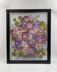 Purple and cool-toned pressed flowers inside two glass panes in a black wood frame